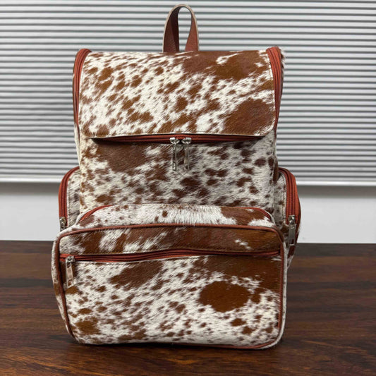 A brown and white cowhide backpack with reddish-brown leather trim, shown standing on a dark wooden surface in front of a window with horizontal blinds.