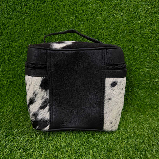 A small black and black-and-white cowhide pouch with a top handle, photographed on a green grass-like surface.