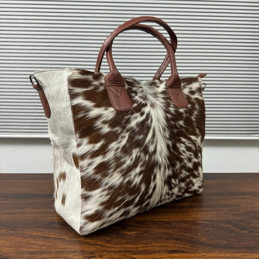 A cowhide tote bag with brown leather handles, resting on a dark wooden table in front of a gray slatted window.