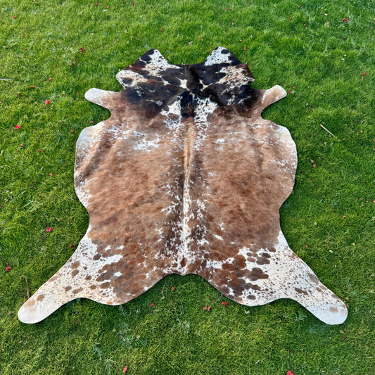 A natural cowhide rug with brown, black, and white mottled patches laid flat on a green grass lawn.