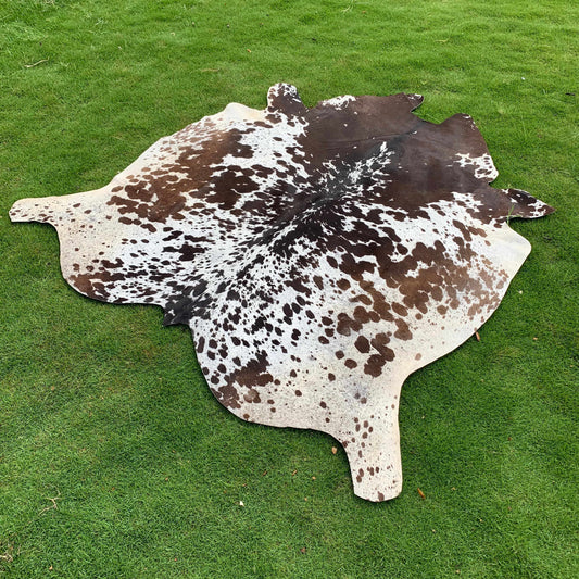 A natural cowhide rug with a brown and white spotted pattern, laid flat on a green grass lawn.