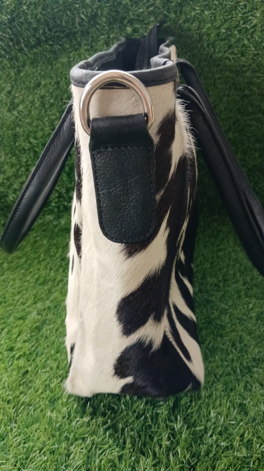A cylindrical shoulder bag with a black and white cow print body, black leather trim, and black handles, shown against a green grass background.