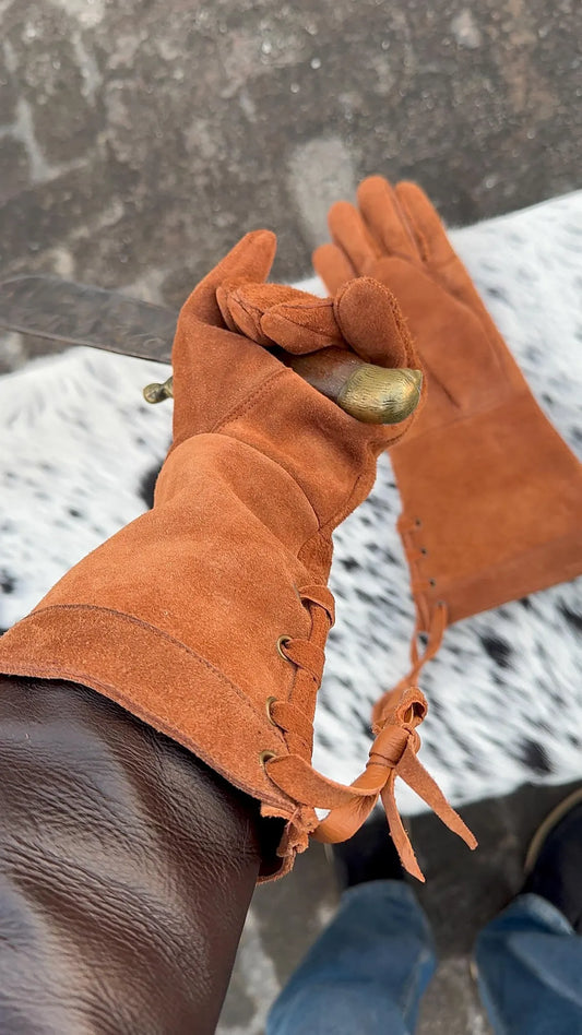 A close-up of a person wearing a pair of long, brown leather work gloves with lace-up cuffs, holding a knife over a snowy ground. The person's jeans are visible at the bottom.
