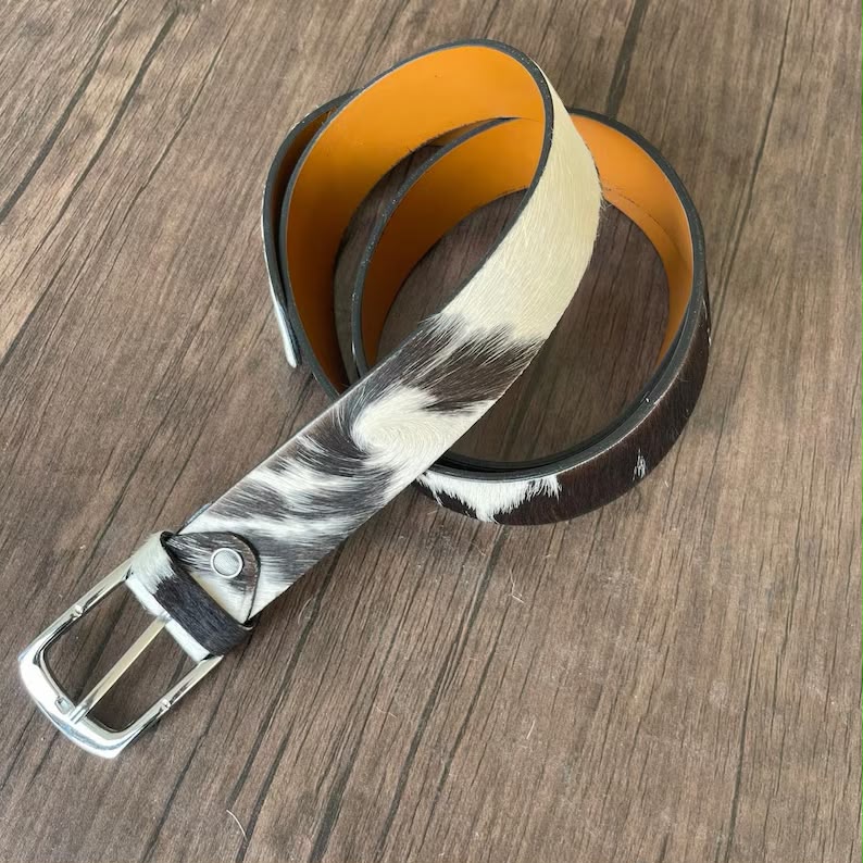 A coiled belt with a black and white cowhide pattern, a bright orange interior lining, and a silver-tone metal buckle, displayed on a brown wood-grain surface.