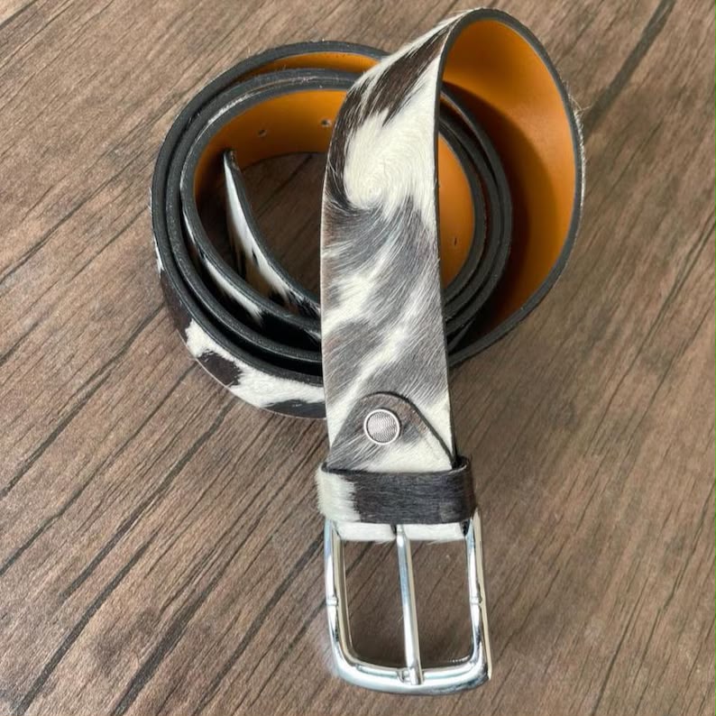 A rolled black and white cowhide belt with a silver buckle, displayed on a brown wooden surface.