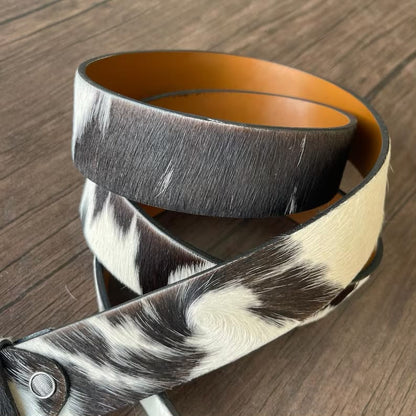 A leather belt with a black and white cowhide pattern and a tan interior, coiled on a dark wood surface.