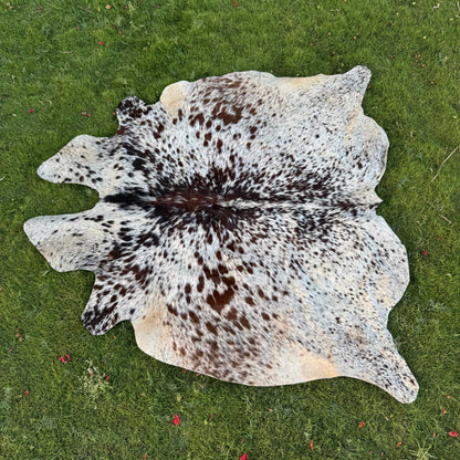 A natural cowhide rug with a brown and black spotted pattern on a white base, laid flat on a green grass lawn.
