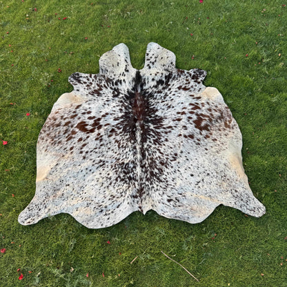 A natural cowhide rug with a black-and-white spotted pattern and tan edges, laid flat on a green grass lawn.