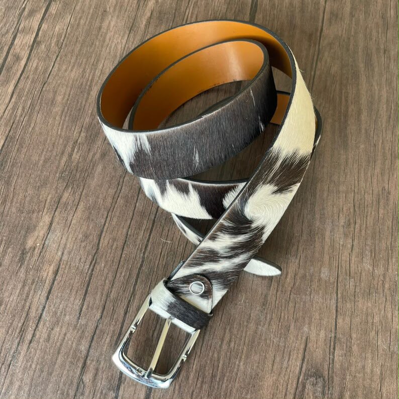 A western-style belt with a black and white cowhide pattern and a tan interior lining, coiled on a brown wood background.