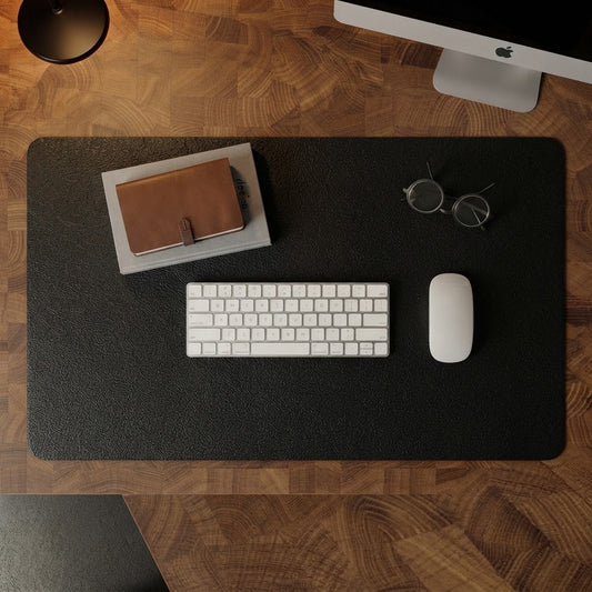 A large black desk mat with a textured surface is laid out on a wooden desk, styled with a white keyboard, white mouse, eyeglasses, and a notebook. An Apple iMac is partially visible in the background.