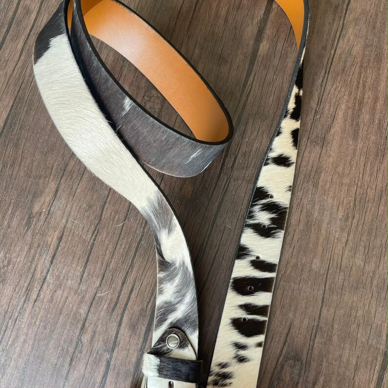A western-style belt with a cowhide pattern, featuring a black and white hair-on-hide strap and a tan leather underside, displayed on a dark wood background.