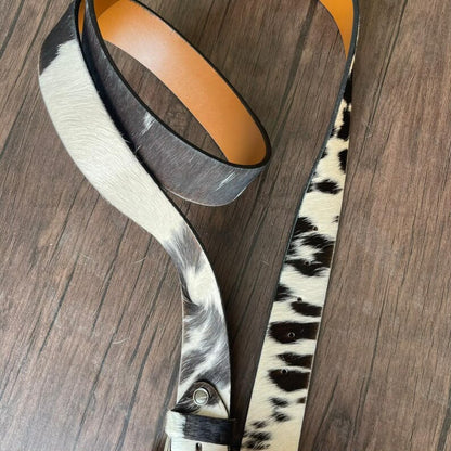 A western-style belt with a cowhide pattern, featuring a black and white hair-on-hide strap and a tan leather underside, displayed on a dark wood background.