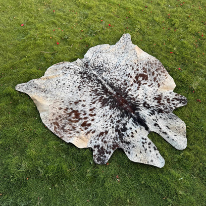 A natural cowhide rug with a black and white spotted pattern and tan edges, laid flat on a green grass lawn.