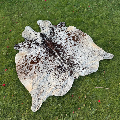 A natural cowhide rug with a brown and black spotted pattern on a cream and light blue base, laid flat on a green grass lawn.