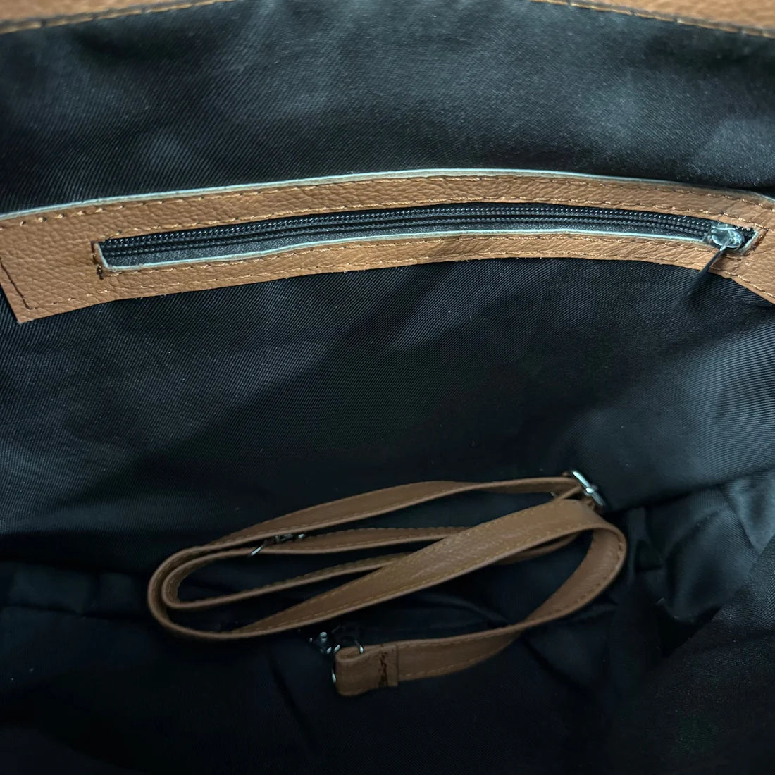 Close-up of the interior of a black fabric bag with a brown leather zipper track and a matching brown detachable shoulder strap coiled inside.