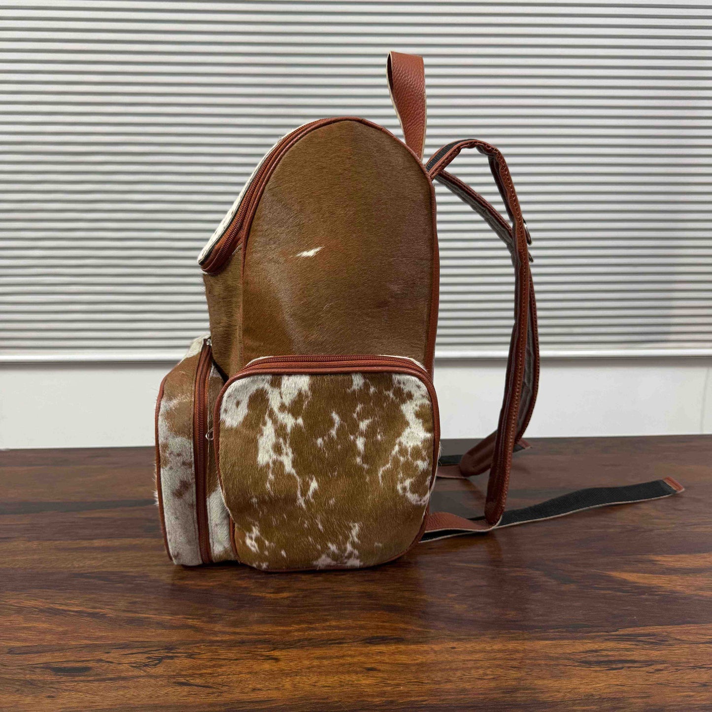 A small backpack made from cowhide, featuring a smooth brown hide on the upper portion and a mottled brown-and-white hide on the front pocket, with reddish-brown leather trim and straps, set on a wooden table against a window blinds background.