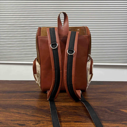 A brown leather backpack shown from the back, featuring black padded shoulder straps with silver metal D-rings, set on a dark wooden table in front of a white slatted window.