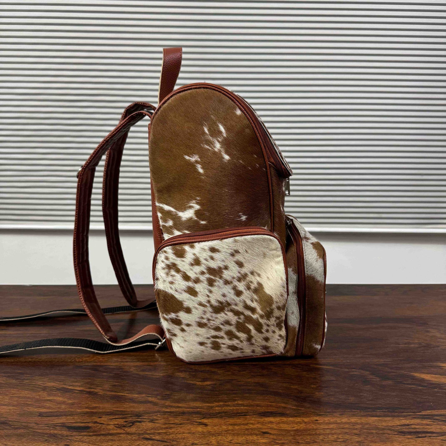 A small backpack with a brown and white cowhide pattern and dark brown leather trim, standing on a wooden table in front of a window with horizontal blinds.