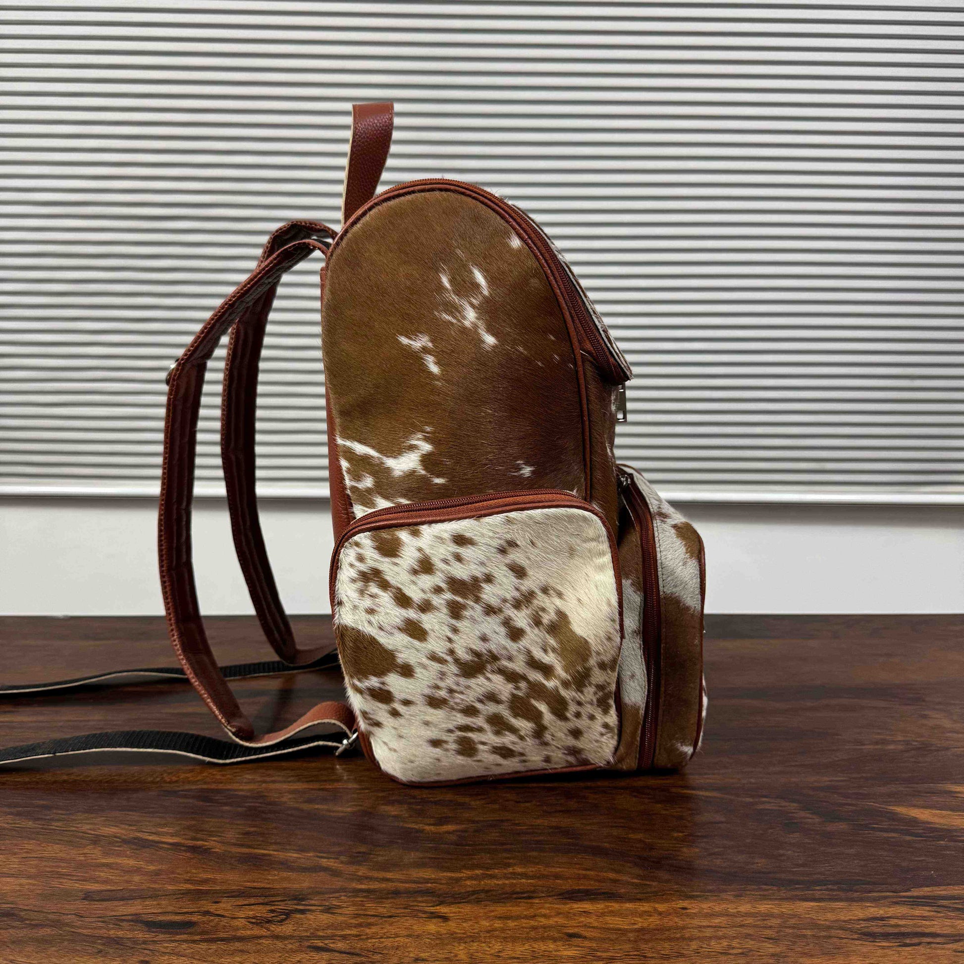 A small backpack with a brown and white cowhide pattern and dark brown leather trim, standing on a wooden table in front of a window with horizontal blinds.