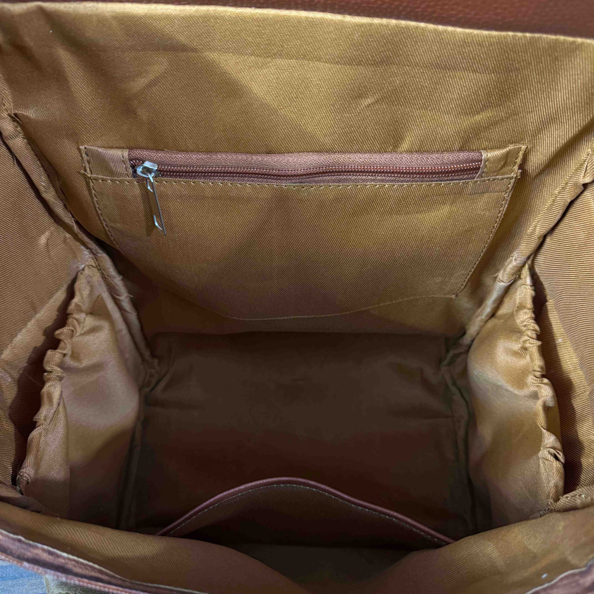Close-up view of the interior lining of a brown bag, showing a tan fabric pocket with a silver zipper pull and gathered fabric sides.