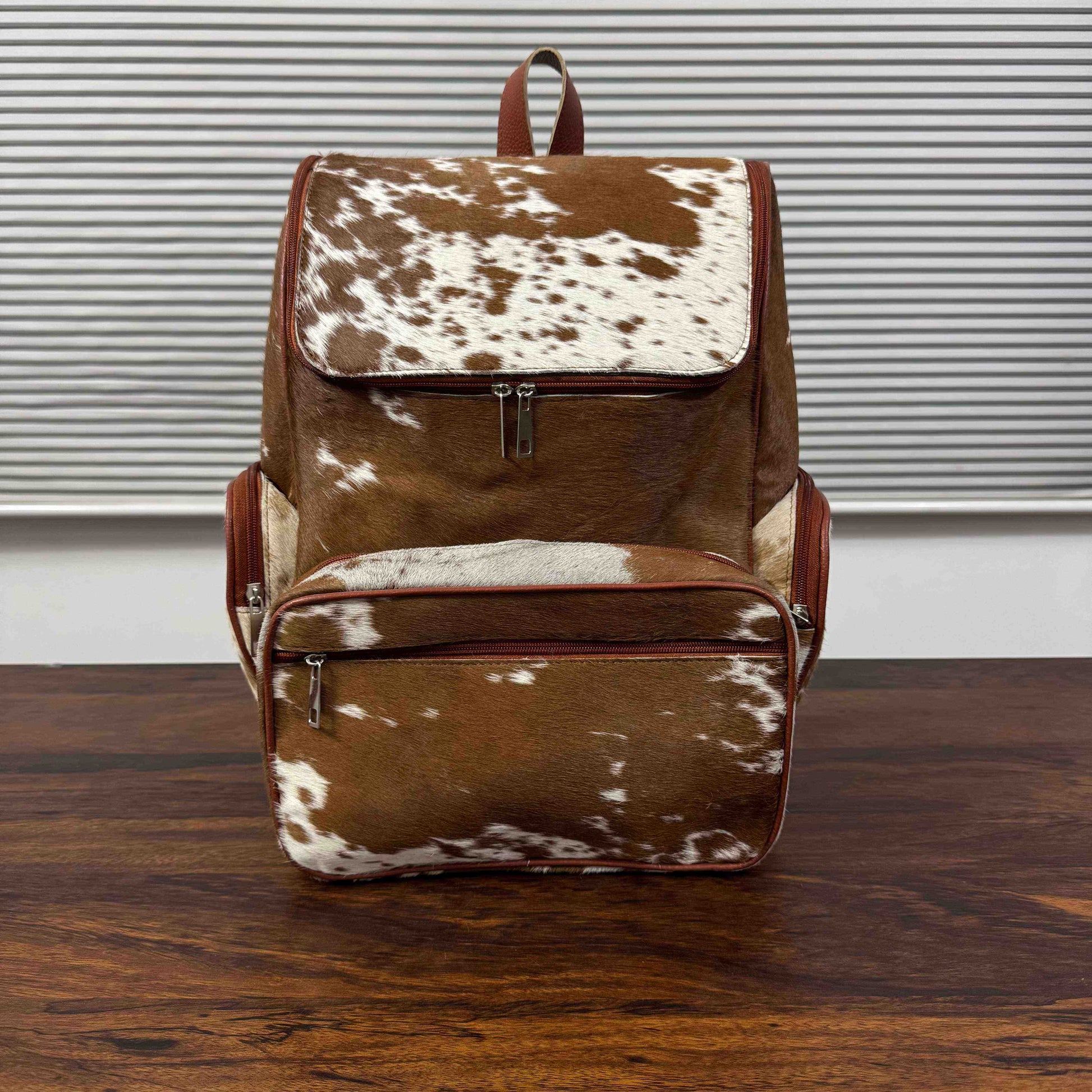 A brown and white cowhide backpack with brown leather trim, standing on a dark wooden table in front of a white wall with horizontal blinds.