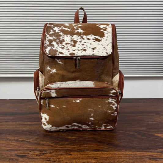 A brown and white cowhide backpack with brown leather trim, standing on a dark wooden table in front of a white wall with horizontal blinds.