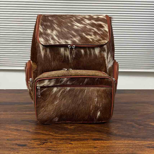 A brown and white cowhide backpack with reddish-brown leather trim, shown standing on a wooden table in front of a window with horizontal blinds.