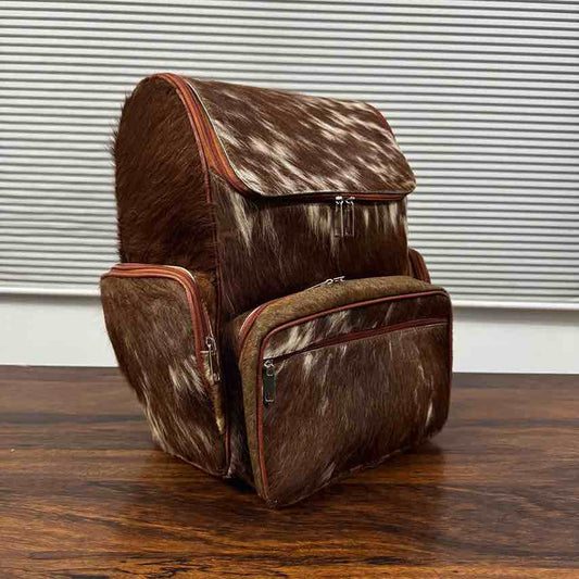 A brown and white cowhide backpack with reddish-brown trim, shown standing on a wooden surface in front of a window with horizontal blinds.