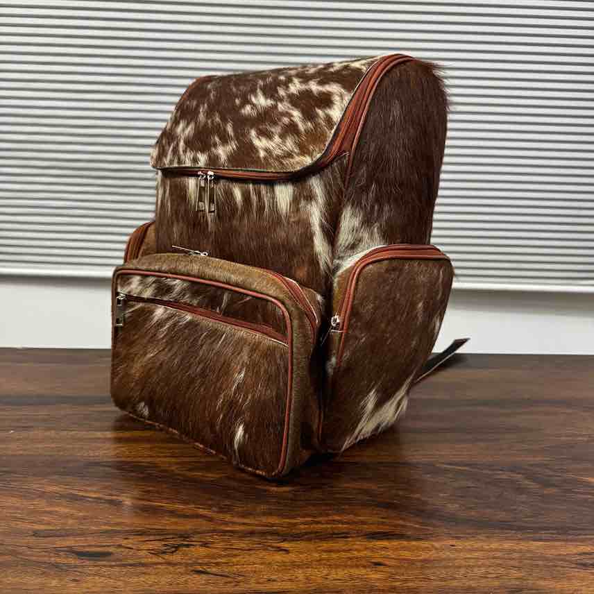 A brown and white cowhide backpack with reddish-brown trim, sitting on a wooden table in front of a window with horizontal blinds.