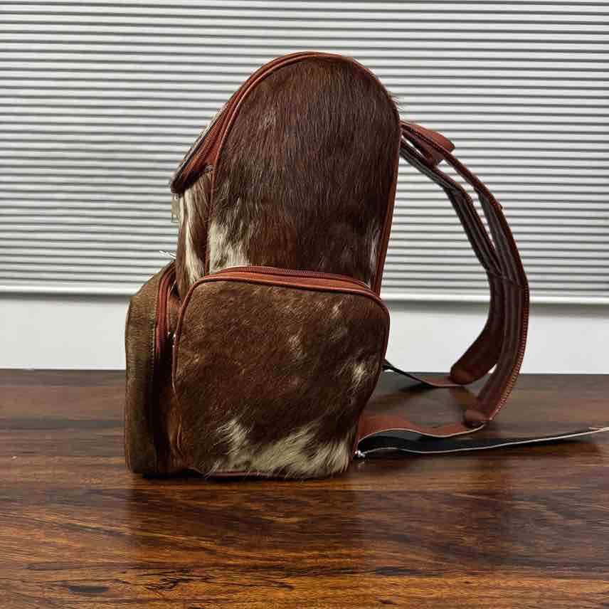 A brown and white cowhide backpack with dark brown leather straps, standing on a dark wooden table in front of a white slatted window.