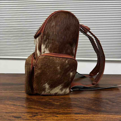 A brown and white cowhide backpack with dark brown leather straps, standing on a dark wooden table in front of a white slatted window.