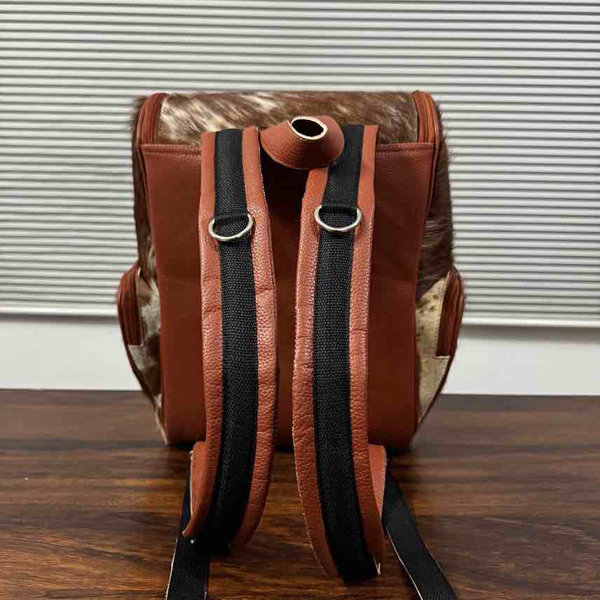 A brown leather and cowhide backpack shown from the back, featuring black padded shoulder straps with silver metal rings, resting on a dark wooden surface against a white slatted window background.