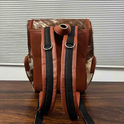 A brown leather and cowhide backpack shown from the back, featuring black padded shoulder straps with silver metal rings, resting on a dark wooden surface against a white slatted window background.