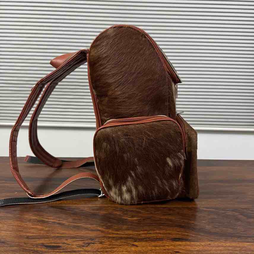 A brown hair-on-hide backpack with matching brown leather straps and trim, resting on a dark wooden table in front of a white slatted window background.