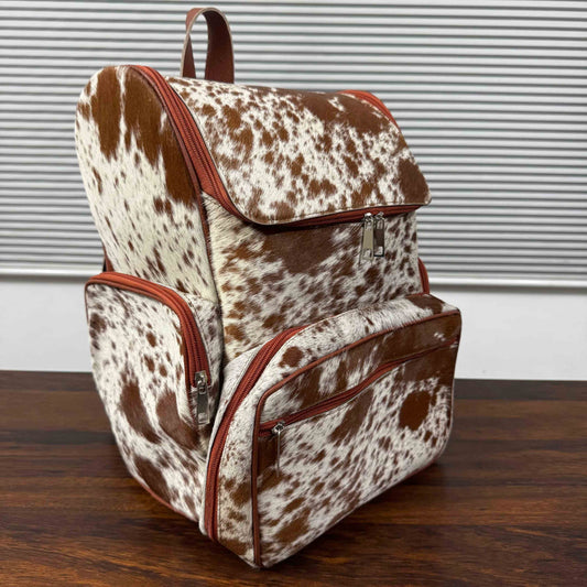 A cowhide backpack with brown and white mottled pattern and reddish-brown trim, standing on a dark wooden table in front of a gray slatted wall.
