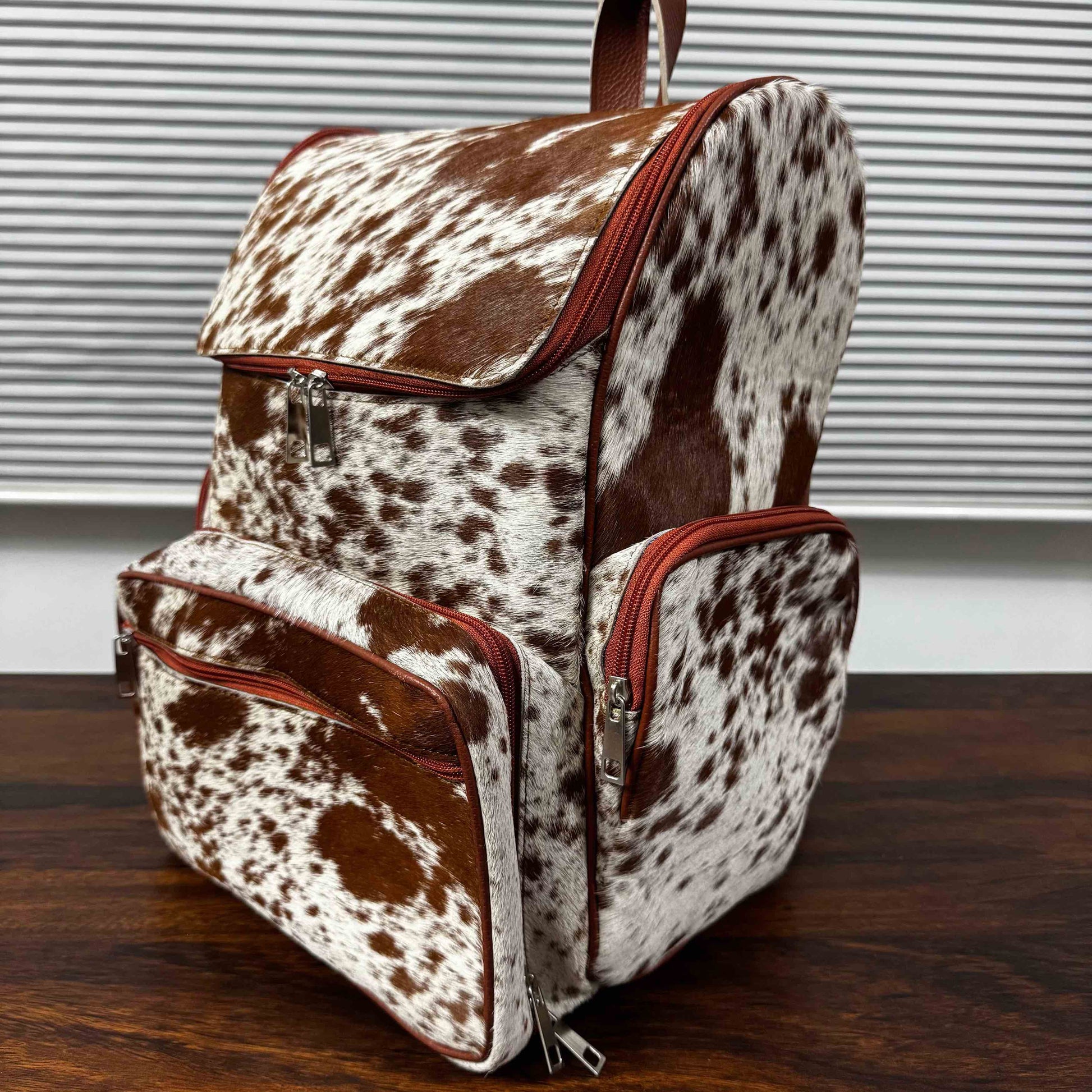 A brown and white cowhide backpack with reddish-brown trim, shown on a dark wooden table in front of a window with horizontal blinds.