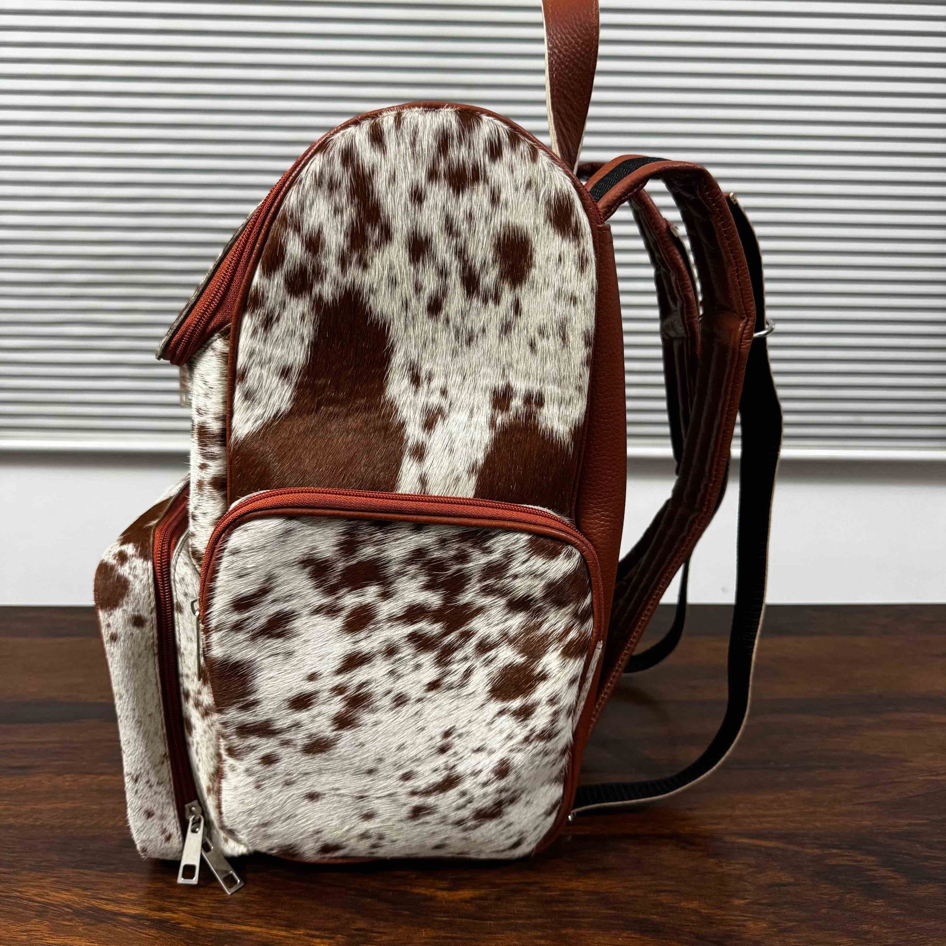A small backpack with a brown and white cowhide pattern, brown faux-leather trim, and black shoulder straps, standing on a dark wooden surface in front of a white slatted window background.