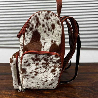 A small backpack with a brown and white cowhide pattern, brown faux-leather trim, and black shoulder straps, standing on a dark wooden surface in front of a white slatted window background.