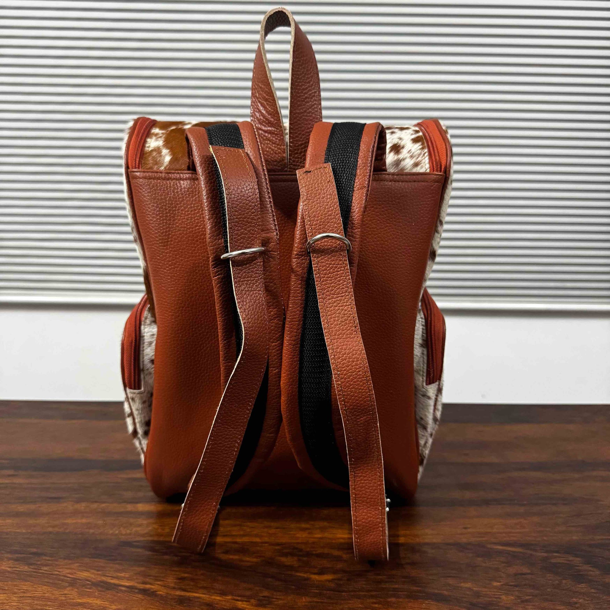 A brown leather backpack with patterned cowhide side panels and adjustable shoulder straps, shown from the back on a wooden surface against a window blinds background.