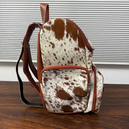A small backpack with a brown and white cowhide pattern and brown leather trim, standing on a wooden surface in front of a window with horizontal blinds.