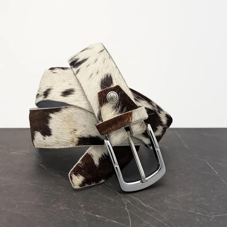A cowhide belt with a brown and white mottled pattern and a silver metal buckle, displayed on a dark gray marble surface against a white background.