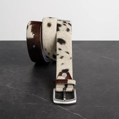 A rolled-up belt with a brown and white cowhide pattern, a dark brown leather end, and a silver-tone metal buckle, displayed on a dark gray marble surface against a light gray background.