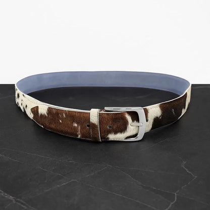 A cowhide belt with a brown and white pattern, a silver buckle, and a light blue interior lining, displayed on a dark gray marble surface against a white background.