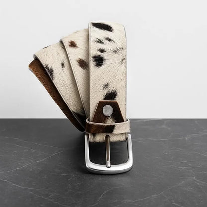 A cowhide belt with a black and white spotted pattern, brown leather trim, and a silver-tone metal buckle, displayed on a dark gray marble surface against a light gray background.