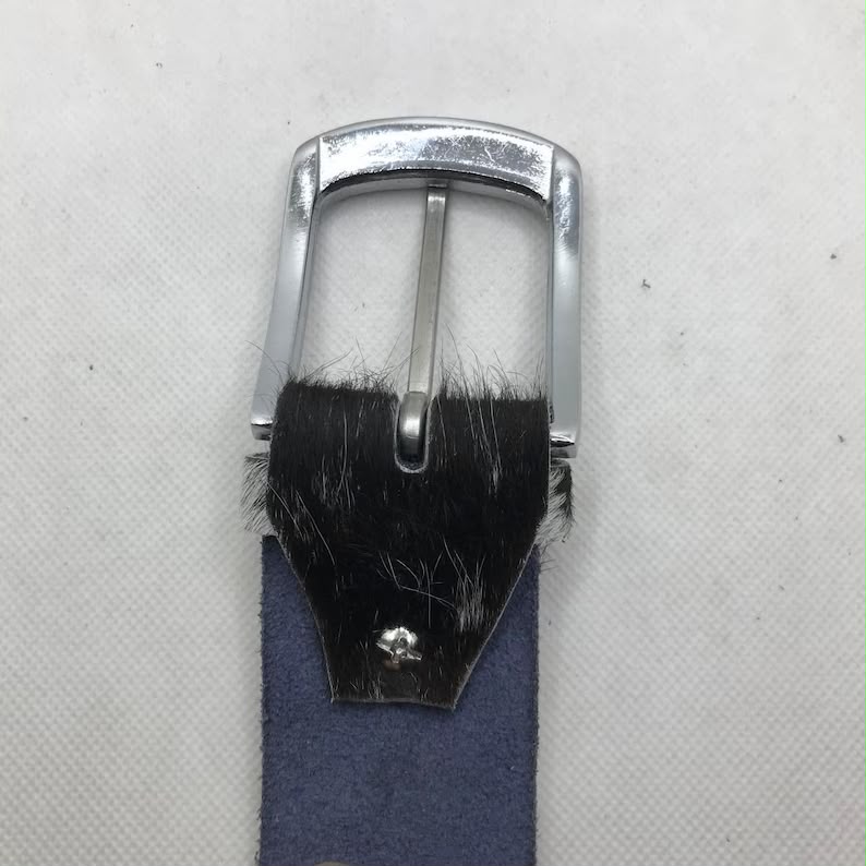 A close-up of a belt buckle with a polished silver-tone metal frame, a black hair-on-hide section, and a blue suede strap on a textured off-white background.