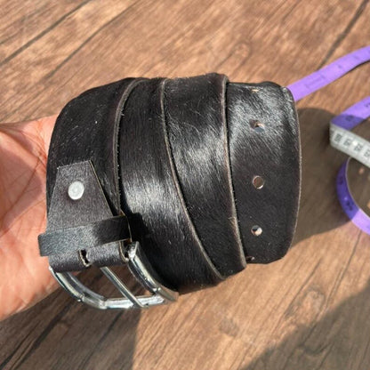 A black cowhide belt with a silver metal buckle, held in a hand over a wooden surface with a purple measuring tape visible in the background.