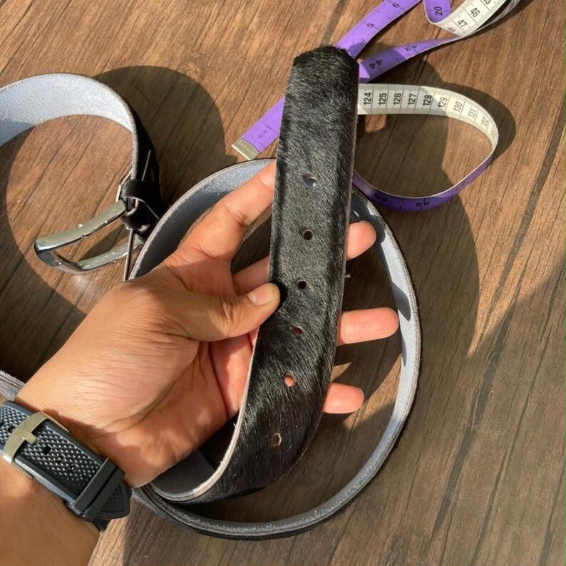 A hand holds a dark brown hair-on-hide belt buckle against a wooden table background, with other belts and a measuring tape visible.