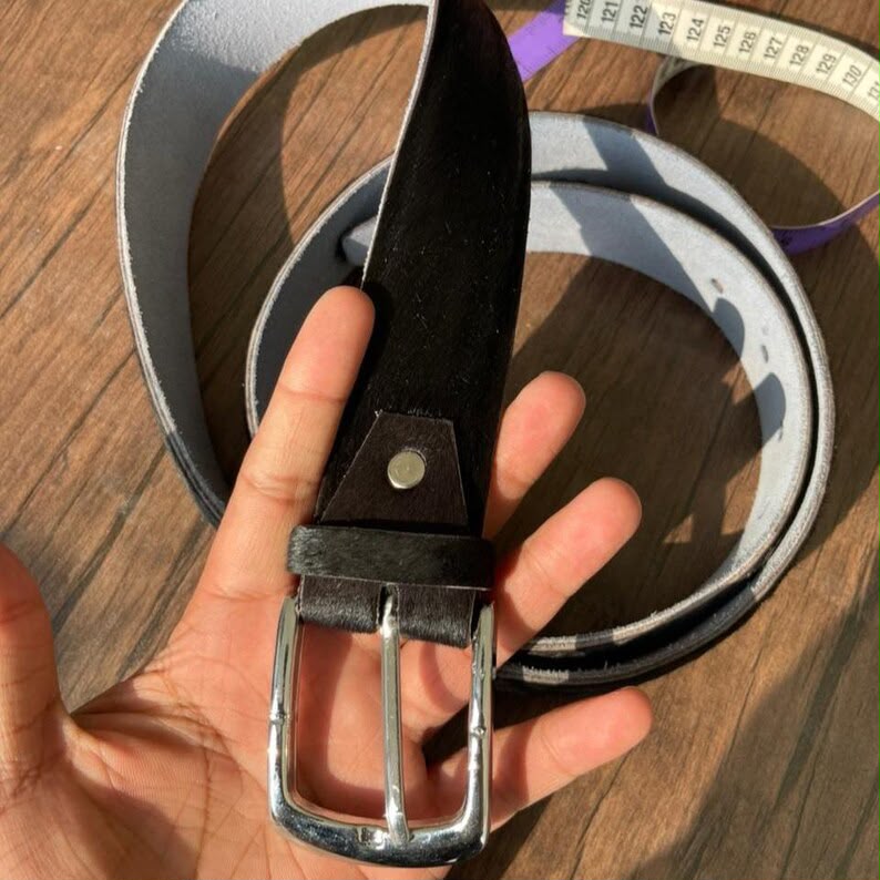 A hand holds a black leather belt with a silver metal buckle, with a coiled light gray belt and a purple measuring tape in the background on a wooden surface.