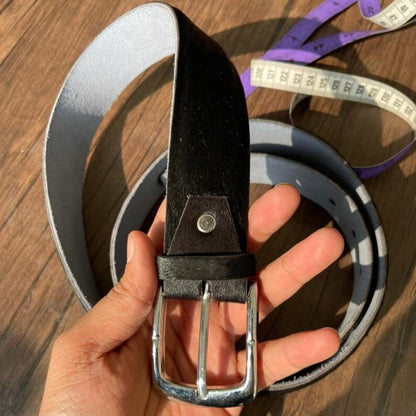 A hand holding a black leather belt with a silver buckle, with a gray belt and a purple measuring tape coiled in the background on a wooden surface.
