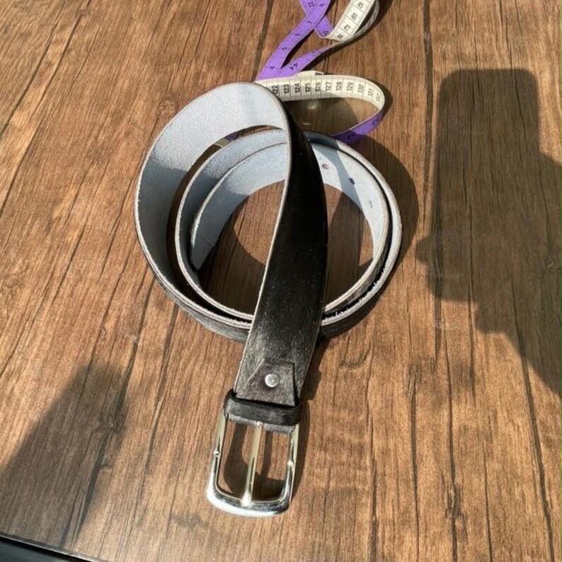 A dark brown leather belt with a silver buckle, coiled on a wooden surface next to a purple measuring tape.